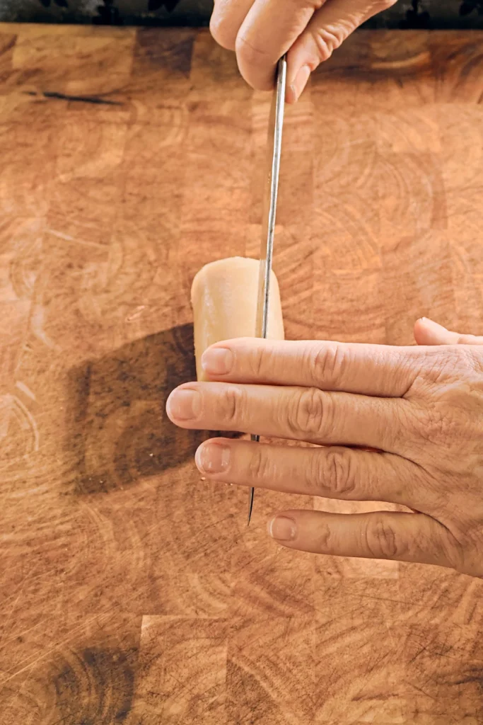 On a large wooden kitchen board lies a roll of marzipan. Two hands are holding a large knife, a start to chop the marzipan.