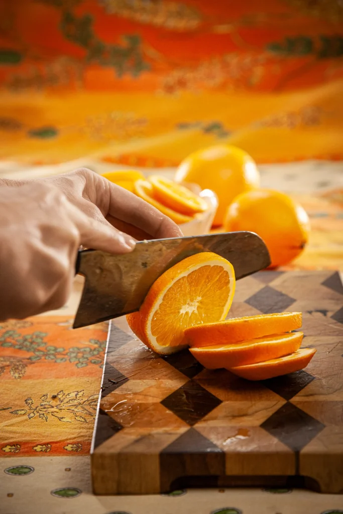 On an patterned orange yellow tablecloth there is a wooden kitchen board. Two hands are holding a large knife and an orange. The knife cuts the orange into slices. Behind the kitchen board is a glass bowl with orange slices and two more oranges.