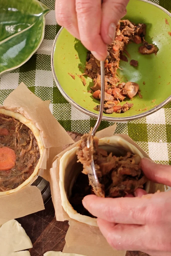 You can see two hands with a spoon filling the dough-lined tins with savoury jackfruit filling. on the table is a green chequered tablecloth.