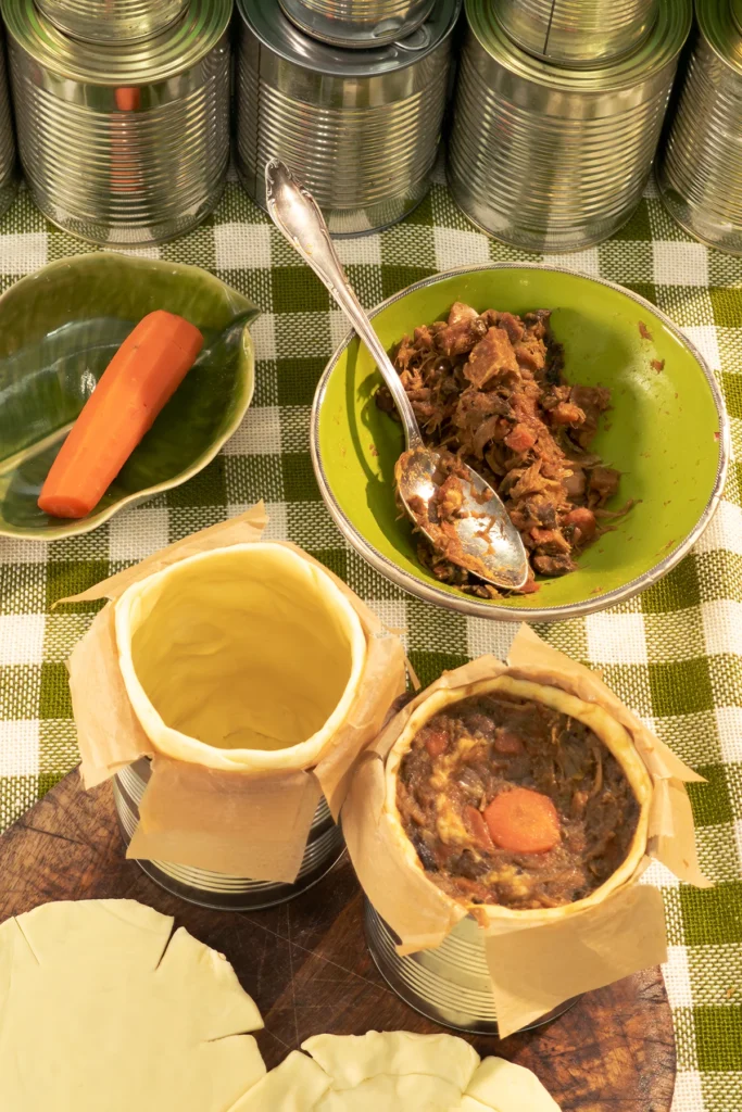 The tins are lined with pastry and one has already been filled with the savoury jackfruit mixture. A cooked carrot has been placed in the centre and savory jackfruit mixture stuffed around it. Behind them is a bowl with savoury jackfruit filling. One cooked carrots is in a shallow bowl. Everything is placed on a green chequered tablecloth. A row of trays can be seen in the background.