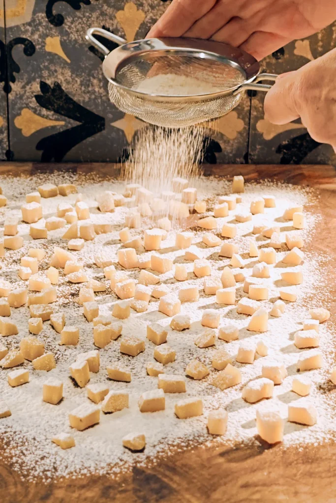 Above a large wooden kitchen board is a hand, holding a sieve with flour, and is flouring the chopped pieces of marzipan that are lying on the wooden surface. There are grey patterned tiles on the wall in the background.