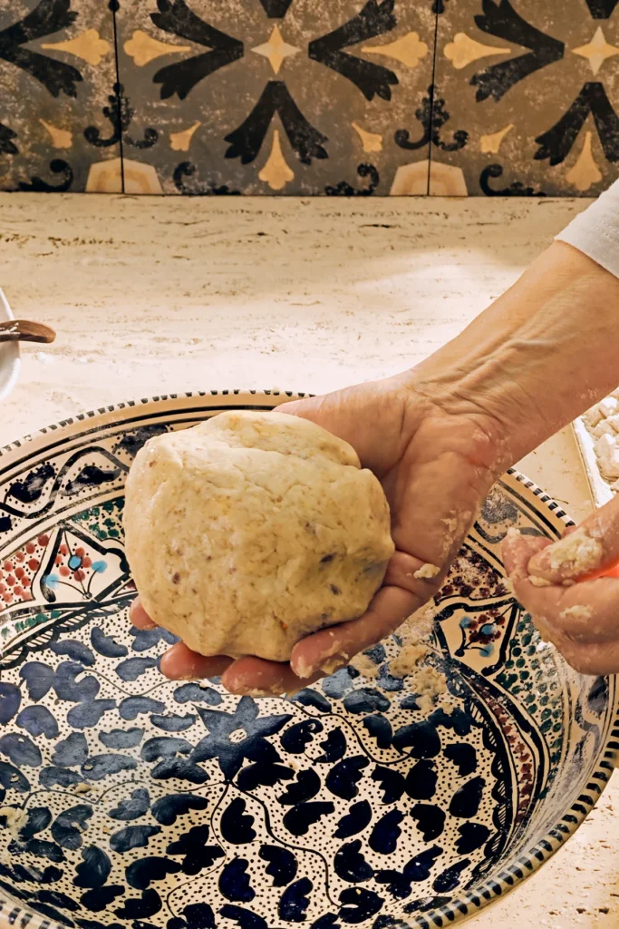 A large patterned ceramic bowl stands on a light-coloured travertine kitchen table with a hand holding the biscotti dough ball. On the wall in the background are grey patterned tiles.