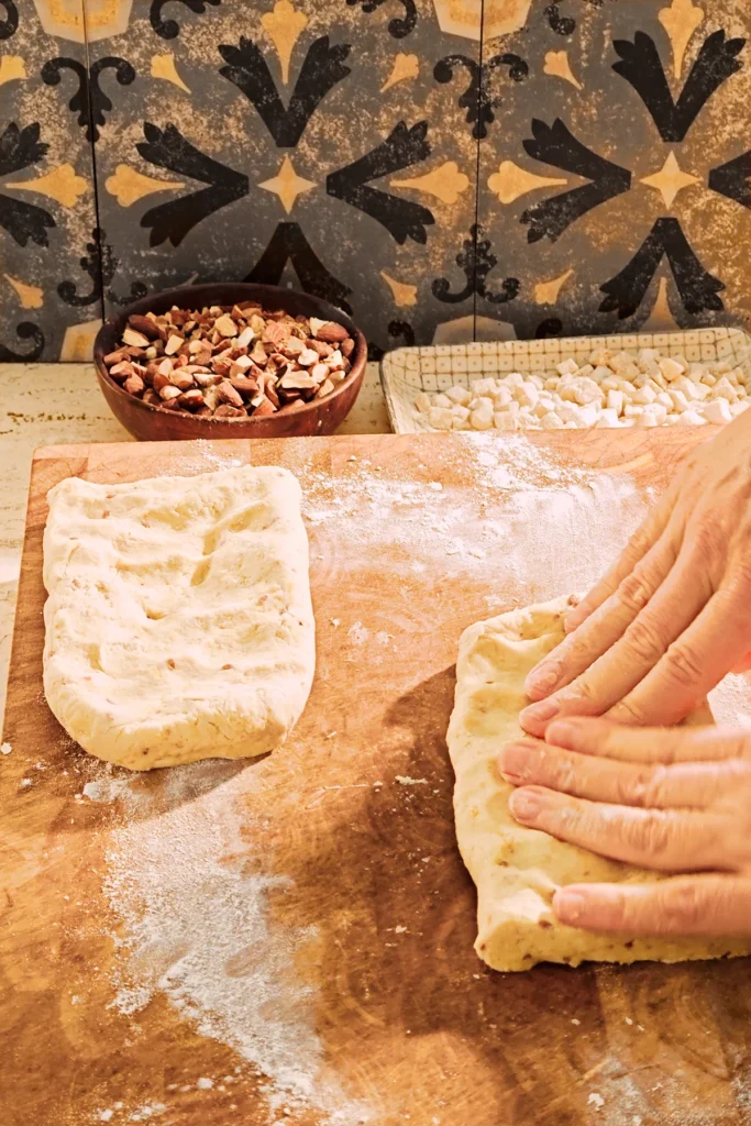 Dough for almond biscotti is kneaded on a large wooden kitchen board. The dough has been divided into two parts. Using two hands, the dough is formed into two rectangular patties about 10mm/0.4 inch thick