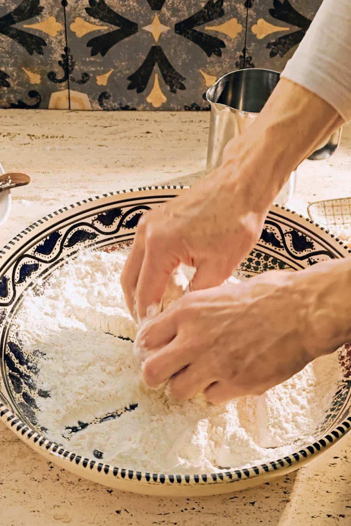 A large patterned ceramic bowl containing flour, starch and baking powder stands on a light-coloured travertine stone kitchen counter. Two hands mix these ingredients together.