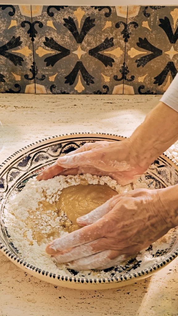 On a light-coloured travertine stone kitchen counter stands a large patterned ceramic bowl of flour, with a mountain of pure apples in the middle of the flour mixture. Two hands begin to mix these ingredients. There are grey patterned tiles on the wall in the background.