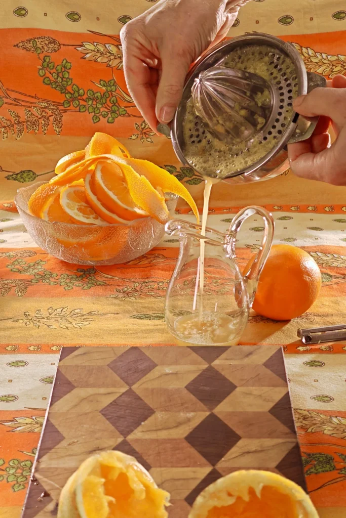 An orange has been squeezed in a citrus juicer. The empty orange peels are placed on a wooden cutting board. Now the fresh orange juice is poured from the press into a glass jug. Behind the jug is a glass bowl with orange slices, all set on a tablecloth in shades of orange with wheat and oat straw motifs.