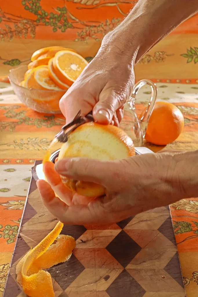 On an orange patterned tablecloth lies a wooden kitchen board with orange peel. Behind it is an empty glass jug and another orange. Two hands are holding an orange that has almost been peeled with the help of a potato peeling knife.