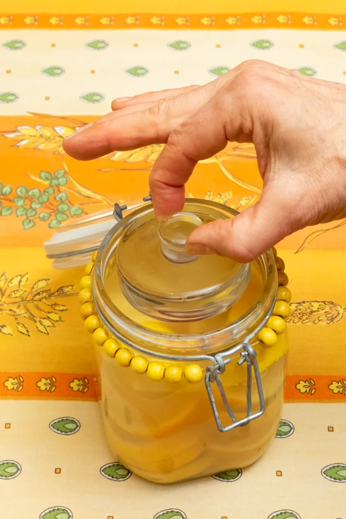 Close-up of an opened preserving jar with preserved lemons. A hand is placing the ferment weight on the lemons. The jar stands on a patterned orange and yellow tablecloth.