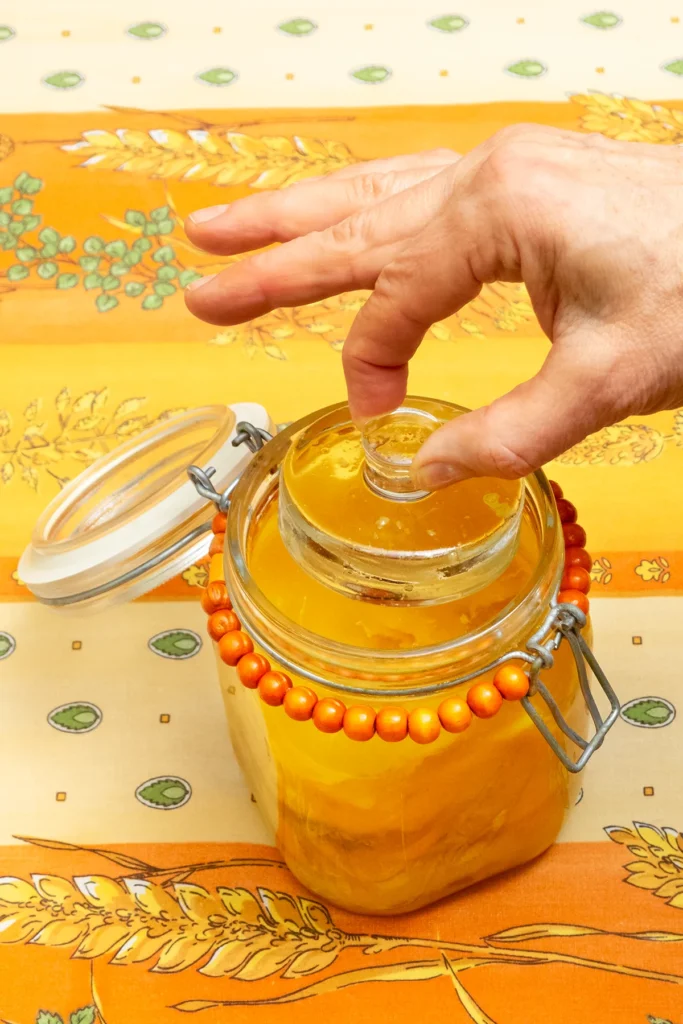 Close-up of an opened preserving jar with preserved oranges. A hand is placing the ferment weight on the lemons. The jar stands on a patterned orange and yellow tablecloth.