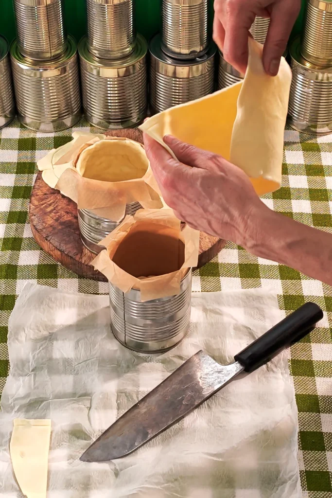 On a green chequered tablecloth lies the baking paper from the quiche dough, with a large knife. The two tin cans lined with baking paper are lined with the cut dough. In the background is a row of tins.