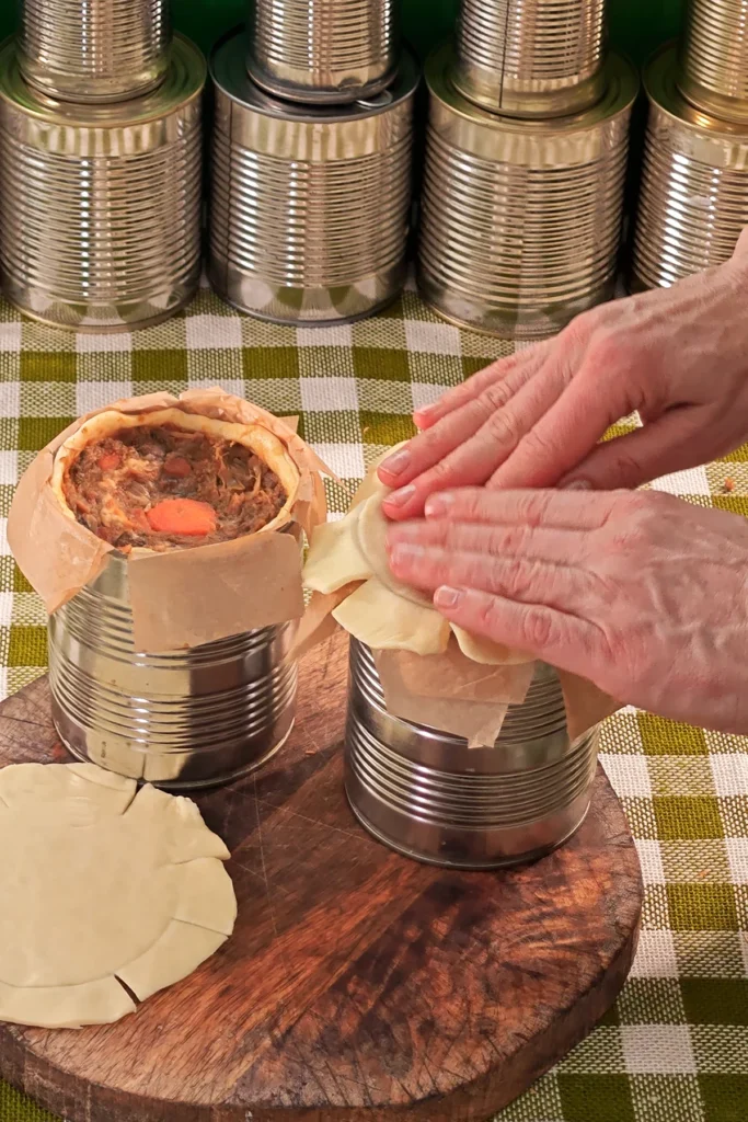 The tin cans are lined with dough and filled with the savoury jackfruit mixture. Before they are turned over to get the pie out of the tin, two hands place a pastry lid on top of the opening. A row of tins stands in the background.