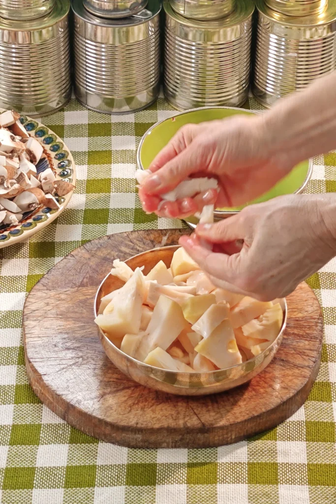 On a green and white chequered tablecloth lies a round wooden kitchen board on which pieces of tinned jackfruit are placed in a silver bowl. Two hands pluck the fibrous pieces of fruit. A row of tins stands in the background.