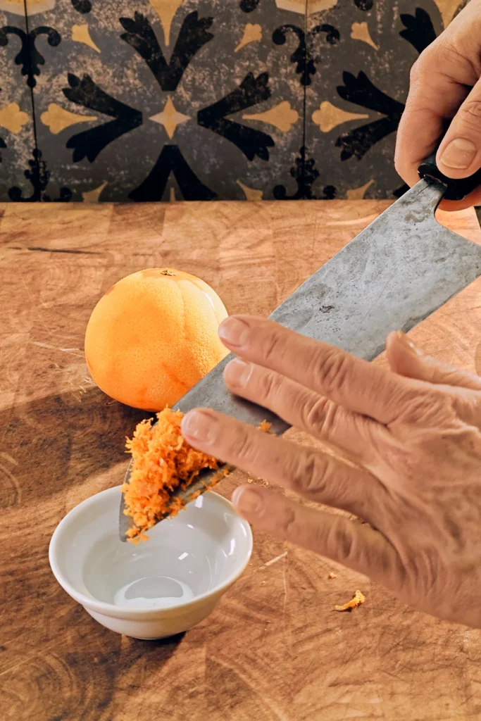 On a large wooden kitchen board stands a small white ceramic bowl, above it a large knife with a mountain of grated orange peel on its blade. One hand wants to push the grated orange into the white bowl. Behind it is a whole orange with grated peel. There are grey patterned tiles on the wall in the background.