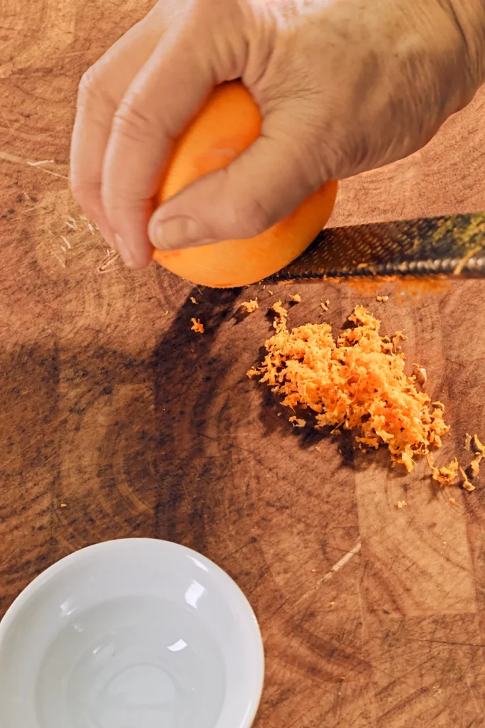 There is a grater on a large wooden kitchen board. One hand holds an orange and rubs it over the grater. There is already a mountain of orange zest under the grater. In front of the picture is a small white bowl.