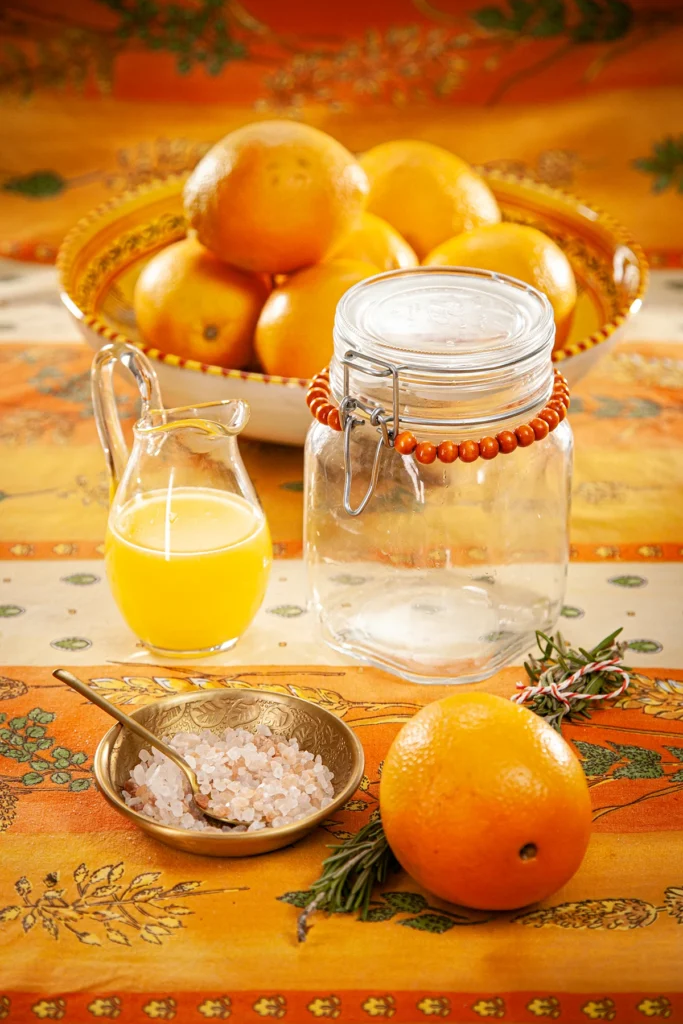 In the background there are oranges in a decorative bowl. In front of it a jug with orange juice, next to a transparent, ready-to-use preserving jar with a string of orange-coloured wooden beads around the lid. In front of the preserving jar is a flat golden bowl with salt and sugar, a bunch of rosemary sprigs and an orange. Everything is laid on an orange-coloured tablecloth with a pattern.