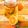 On an orange tablecloth with wheat and oat straw motifs stands a preserving jar with the preserved oranges inside. Behind it is organic orange. In the foreground are two halved peeled oranges peel and a twig of rosmarin.