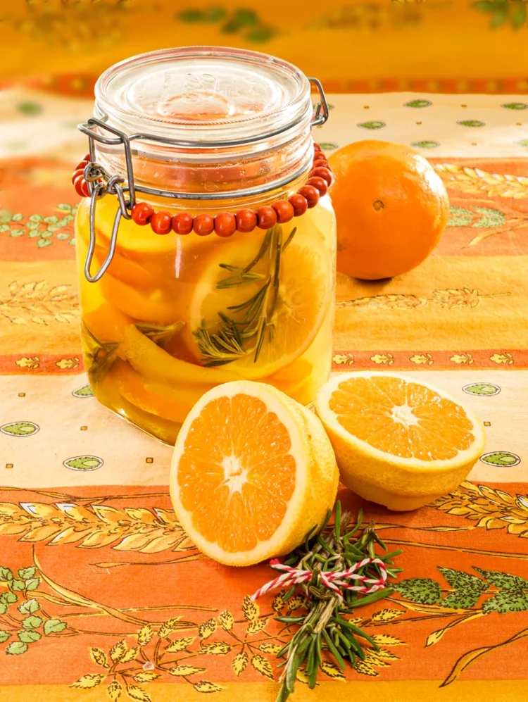 On an orange tablecloth with wheat and oat straw motifs stands a preserving jar with the preserved oranges inside. Behind it is organic orange. In the foreground are two halved peeled oranges peel and a twig of rosmarin.