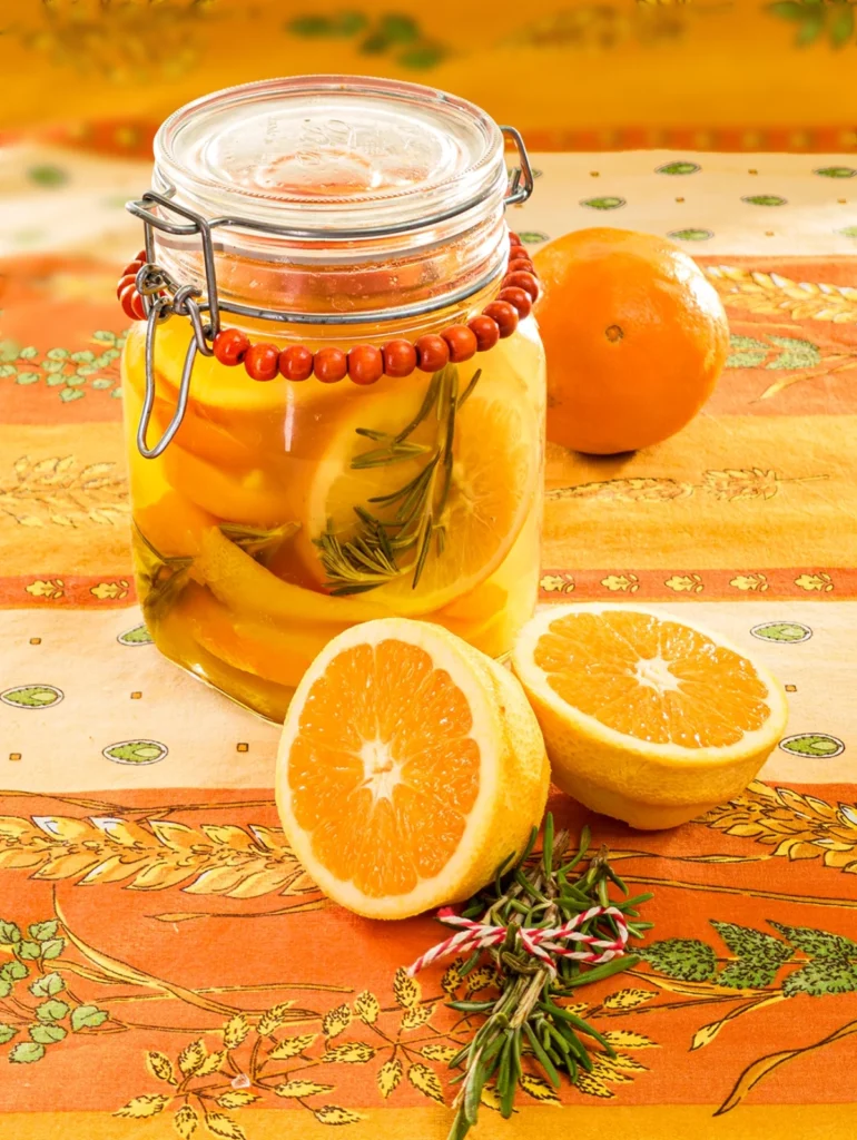 On an orange tablecloth with wheat and oat straw motifs stands a preserving jar with the orange slices inside. Behind it is organic orange. In the foreground are two halved peeled oranges peel and a twig of rosmarin.