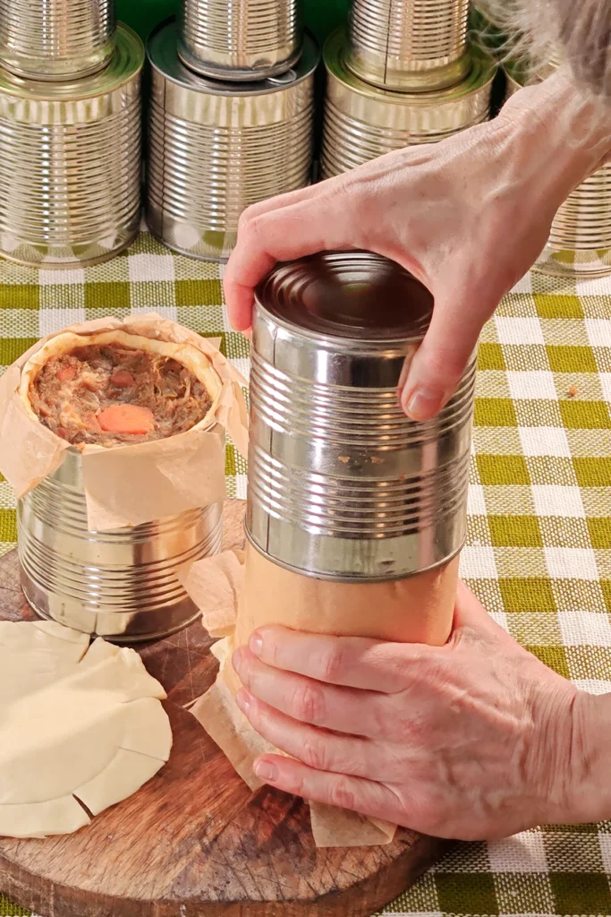 One hand pulls on the tin, the other holds the filled dough roll and pulls it out of the tin by the baking paper. Behind it, another filled pie in a tin, still open, the pastry lid lying in front of it on a wooden kitchen board. A row of tins in the background. Everything is placed on a tablecloth with a green and white check pattern.