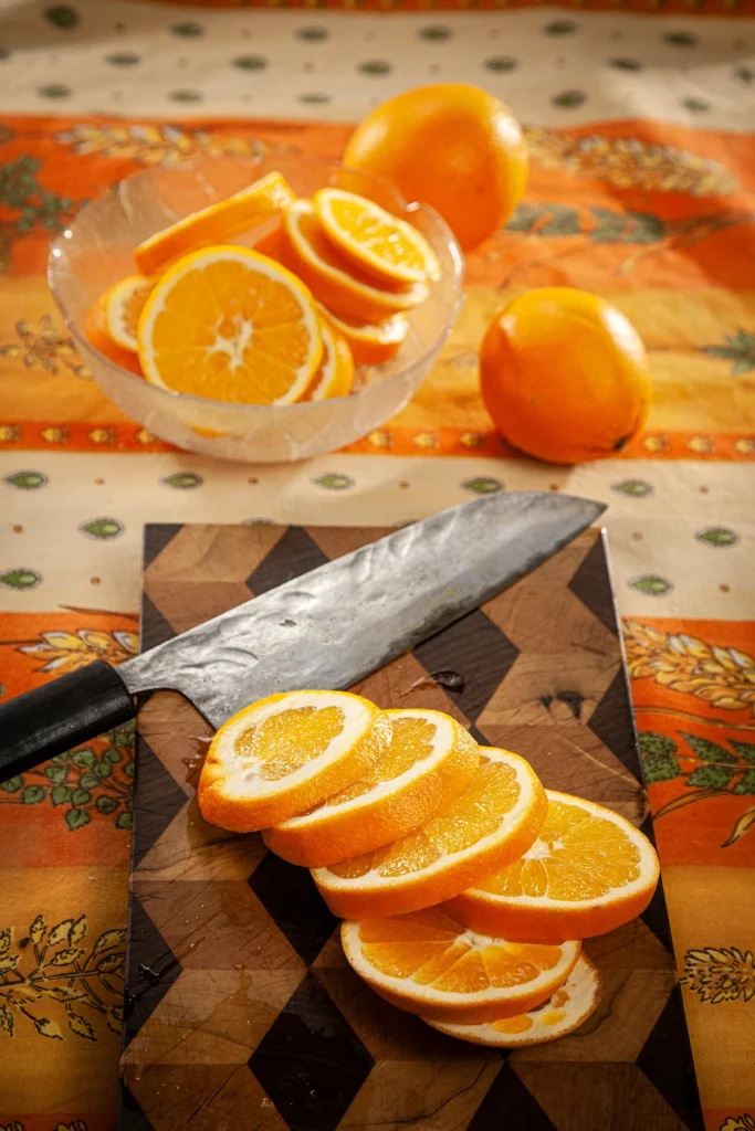 On an patterned orange yellow tablecloth lies a wooden kitchen board with a huge knife and orange slices. Behind the kitchen board is a glass bowl with orange slices.