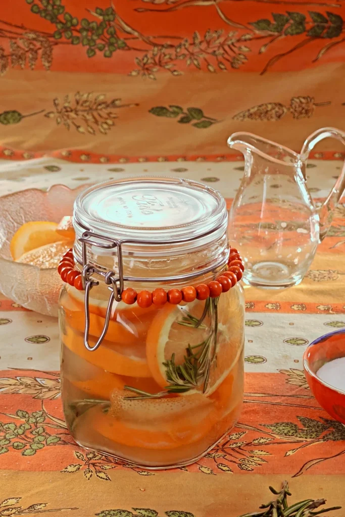 The closed preserving jar with orange slices and herbs is ready. The jar with the preserved oranges is standing on an orange and yellow patterned tablecloth. Behind is a glass bowl with left-over orange slices and an emty jug.