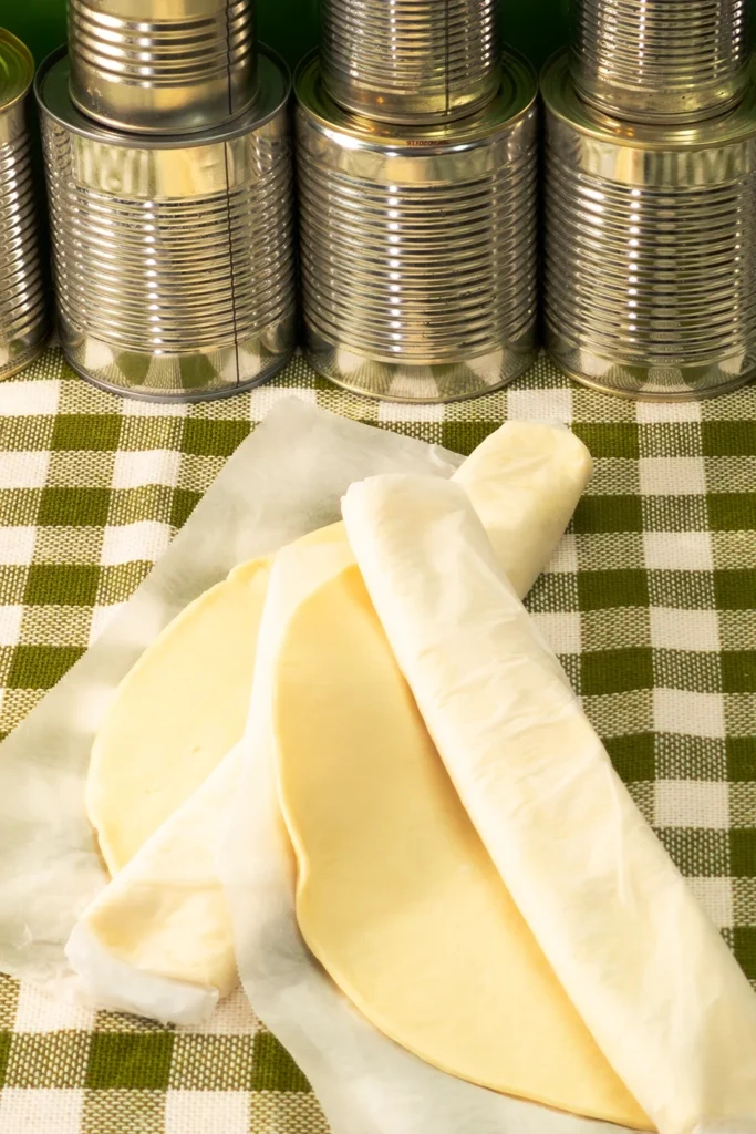 Two rolls of ready-made quiche dough lie on a tablecloth with a green and white check pattern. A row of tins in the background.