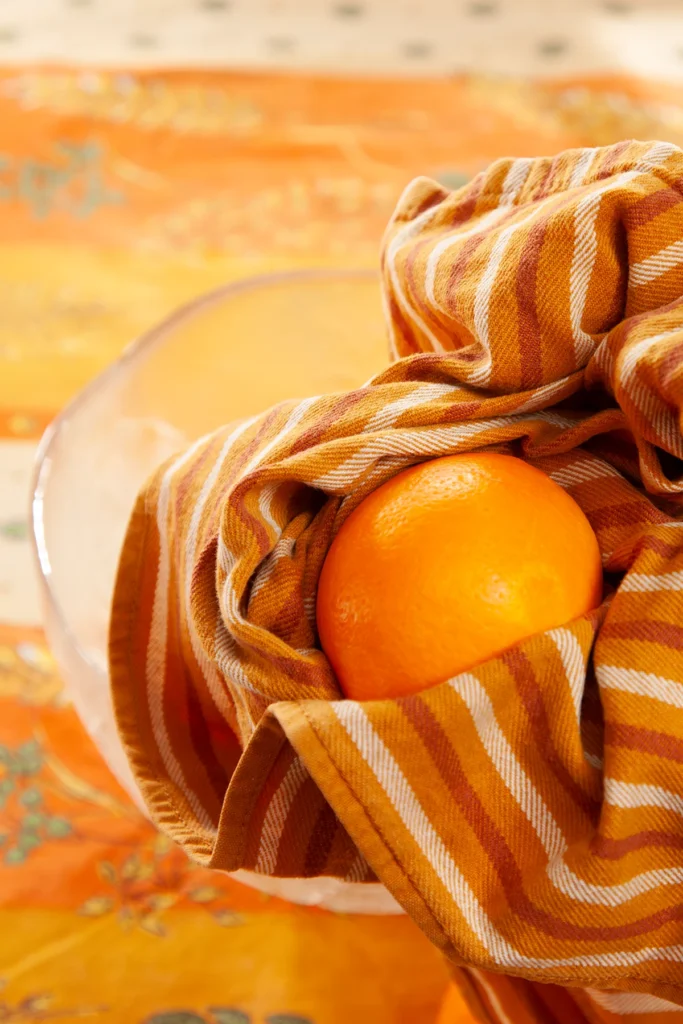 The orange is dried by two hands with a striped kitchen towel. The background is a tablecloth in orange tones with wheat and oat straw motifs.