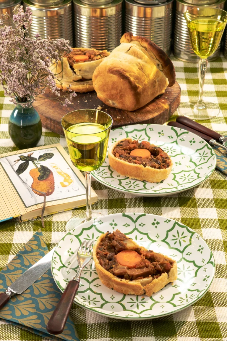 Savory jackfruit pie is served on a beautifully set table with green patterned plates. The pie has a crispy golden crust and a rich, hearty filling with jackfruit, carrots, and mushrooms. Glasses of non-alcoholic wine, fresh herbs, and vintage-style decor create a cozy and inviting atmosphere. Empty, shiny silver tins in the background are reminiscent of the pie's creator, the tin shape.