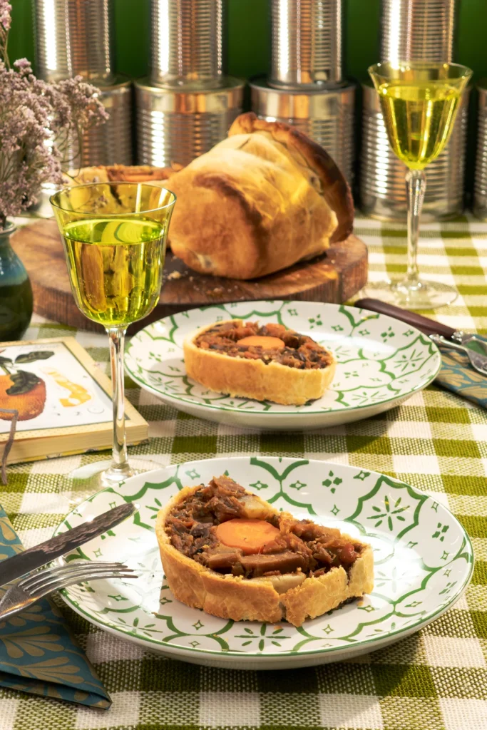 Savory jackfruit pie is served on a beautifully set table with green patterned plates. The pie has a crispy golden crust and a rich, hearty filling with jackfruit, carrots, and mushrooms. Glasses of non-alcoholic wine, fresh herbs, and vintage-style decor create a cozy and inviting atmosphere. Empty, shiny silver tins in the background are reminiscent of the pie's creator, the tin shape.