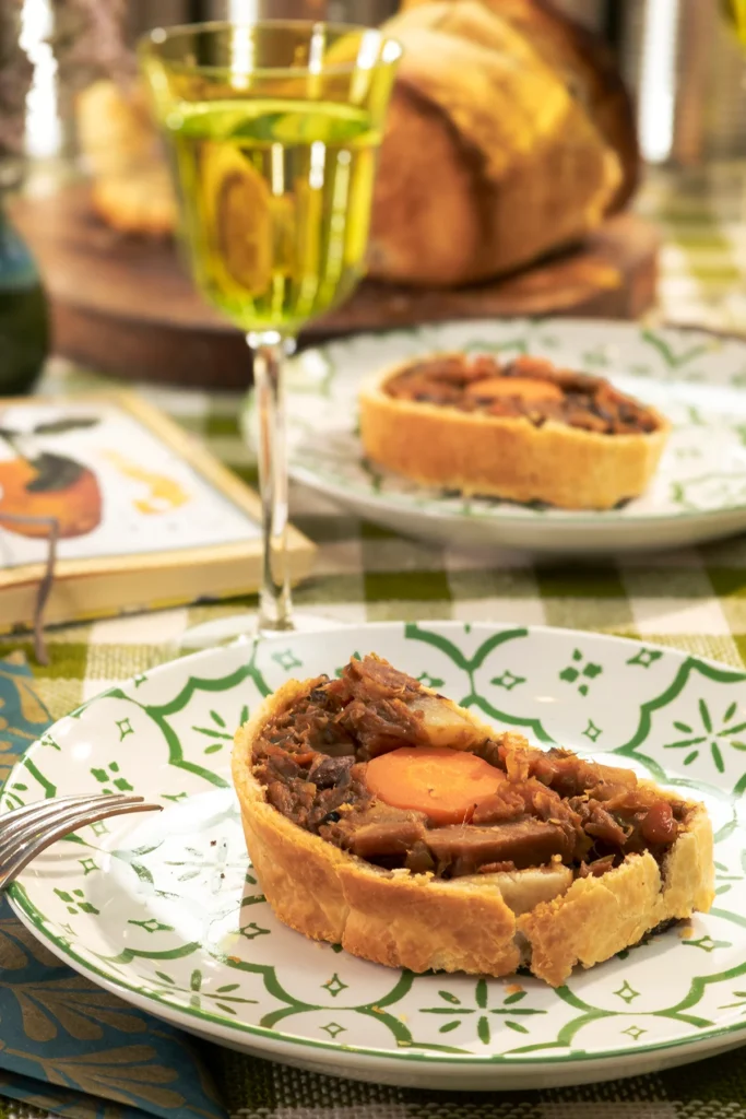 Close view on savory jackfruit pie, that is served on a beautifully set table with green patterned plates. The pie has a crispy golden crust and a rich, hearty filling with jackfruit, carrots, and mushrooms. Glasses of non-alcoholic wine, fresh herbs, and vintage-style decor create a cozy and inviting atmosphere.