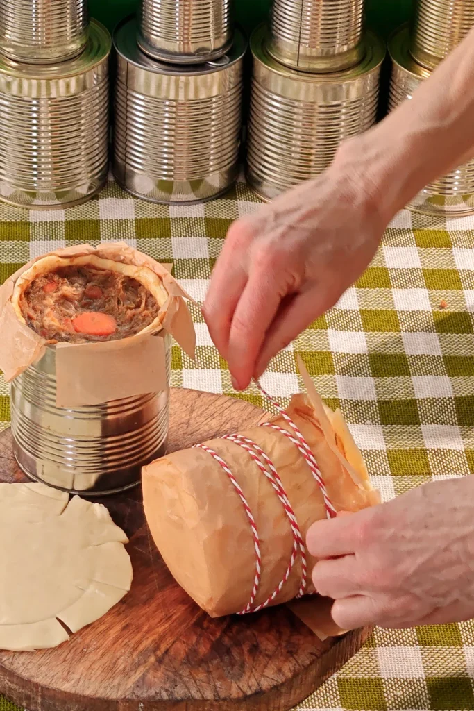 Hands fix the baking paper around a roll of dough with a heat-resistant cord. Behind it, another filled savoury jackfruit pie in a still open tin, the pastry lid resting in front of it on a wooden kitchen board. A row of tins in the background. Everything is placed on a tablecloth with a green and white chequered pattern.