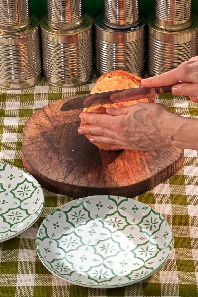 On a green and white chequered tablecloth lies a round wooden kitchen board with a savoury jackfruit pie on it. Two hands cut off the first slice with a serrated knife. In front of them are two patterned plates.