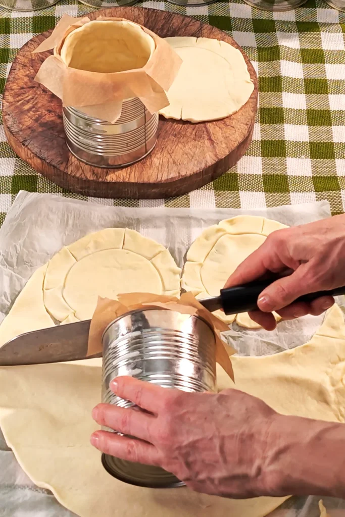 The tins are lined with dough. The dough is rolled out on baking paper and a hand with a knife cuts the dough to size. Another hand holds the tin to take measurements. The base is a green chequered tablecloth.