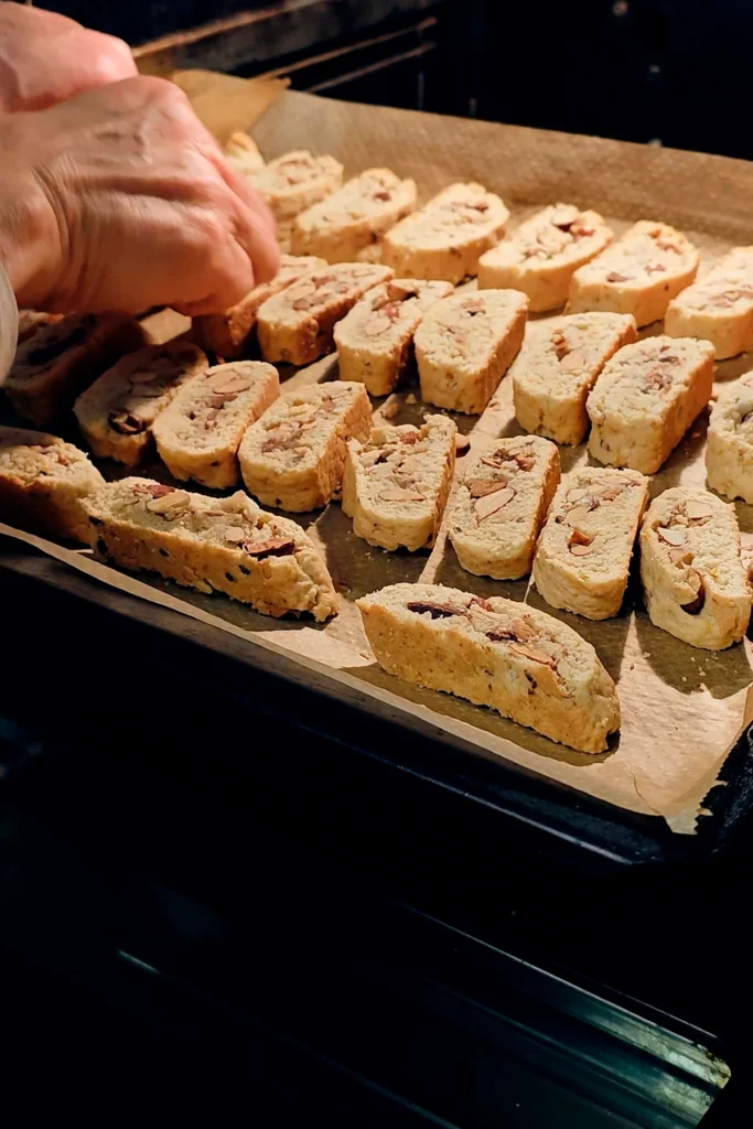 A hand is on the open oven on the baking tray with the slices of vegan almond biscotti with orange and marzipan, turning them for the last baking cycle.