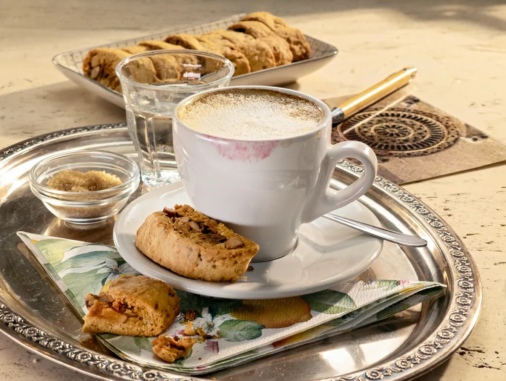 A cozy coffee moment with vegan almond biscotti with orange and marzipan. A cappuccino with frothy milk sits on a silver tray, paired with a crunchy biscotti filled with almonds and marzipan. A plate of freshly baked biscotti is in the background, creating a warm and inviting atmosphere.