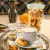 A cosy coffee moment with vegan almond biscotti with orange and marzipan. A cappuccino with milk foam stands on a silver tray, paired with a crispy biscotti with an almond and marzipan filling. In the background is a glass of freshly baked biscotti, next to it is an orange, an Italian travel guide, a vase of white tulips, a pen and postcards. On the wall in the background are grey patterned tiles.