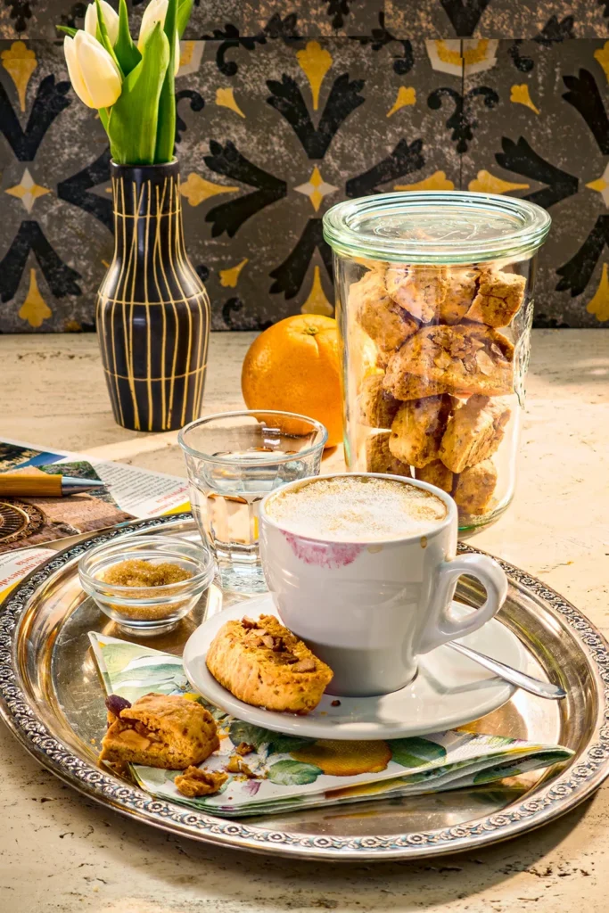 A cosy coffee moment with vegan almond biscotti with orange and marzipan. A cappuccino with milk foam stands on a silver tray, paired with a crispy biscotti with an almond and marzipan filling. In the background is a glass of freshly baked biscotti, next to it is an orange, an Italian travel guide, a vase of white tulips, a pen and postcards. On the wall in the background are grey patterned tiles.