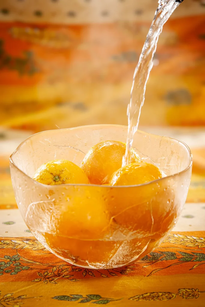 Oranges in a glass bowl are doused with hot water. The bowl stands on a patterned orange yellow tablecloth.