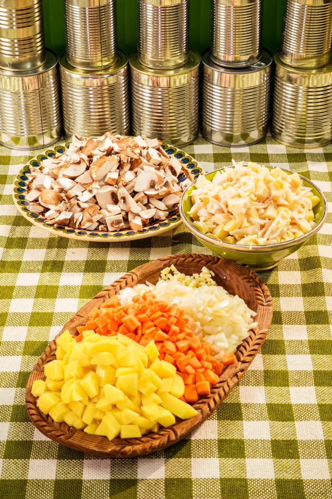 On a tablecloth with a green and white chequered pattern ist a wooden plate in front with the finely cut vegetables. Behind is a bowl with the jackfruit and a plate with the chopped cremini mushrooms. In the background is a row of tins.