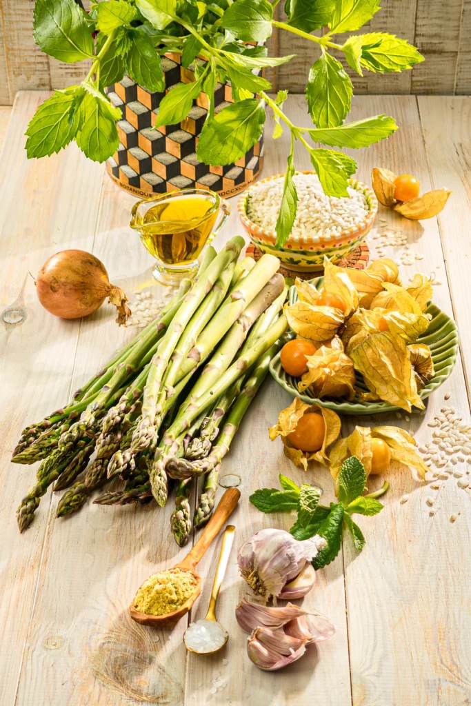 On a wooden table are the ingredients for one-pot asparagus risotto: fresh green asparagus, risotto rice, physalis, mint, garlic, olive oil, an onion and spices.