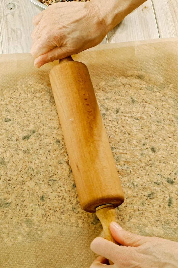 Two hands hold a rolling pin and roll the crispbread dough out between two baking papers.