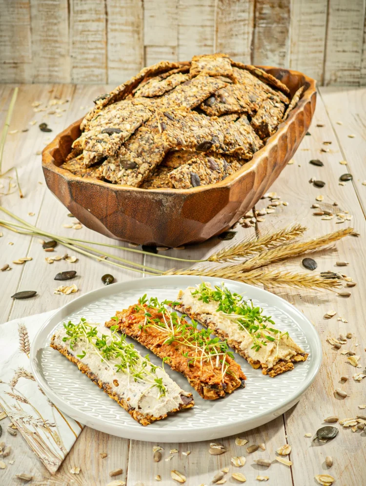 Homemade crispbread with seeds served in a wooden bowl and on a plate topped with various spreads and fresh sprouts.