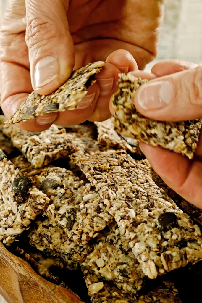 Two hands break the baked crispbread with seeds. Below is a wooden bowl filled with crispbread.