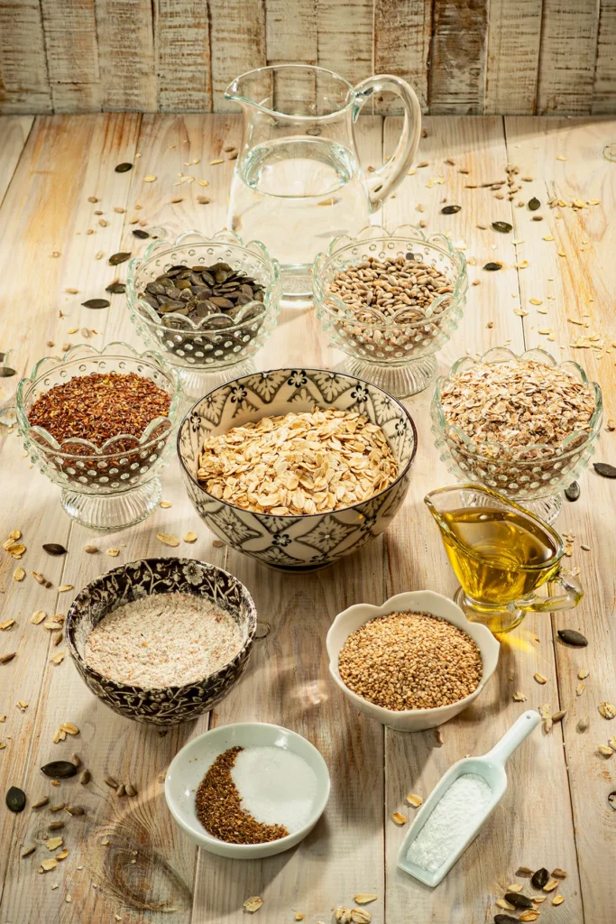 Ingredients for easy crispbread with seeds, including oat and spelt flakes, seeds, flour, oil, and water, arranged in bowls on a wooden surface.