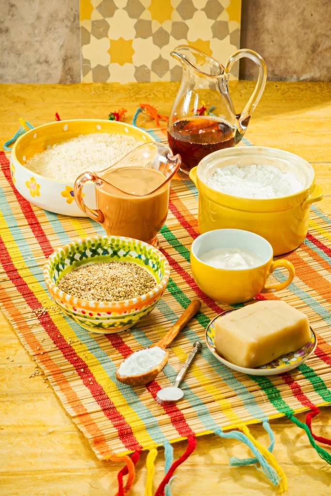 Ingredients for sesame cookies with ground almonds, tahini, maple syrup, cornstarch, soy yogurt, marzipan and sesame seeds arranged in bowls and jugs on a colorful raffia mat.
