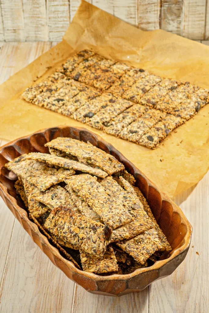 A wooden bowl full of crispbread with seeds and on baking paper a ready-baked plate of crispbread.