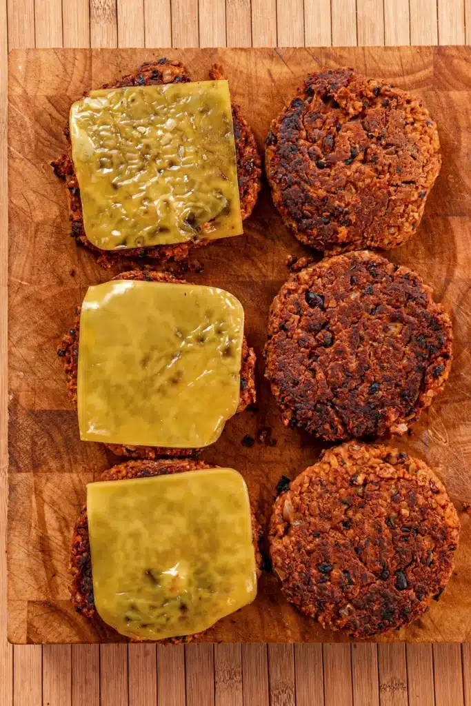 Six BBQ tempeh burger patties are seared, three are topped with vegan cheese.